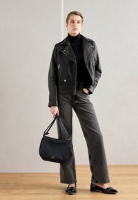 Giacca da moto in pelle nera sopra un dolcevita nero, abbinata a pantaloni a gamba larga grigi e ballerine nere. Tiene in mano una borsa nera con dettagli dorati.