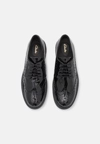 Schwarze Lacklederschuhe mit Brogue-Verzierungen, Schnürverschluss, glatter Textur und runder Zehenform. Branding auf der Innensohle sichtbar.