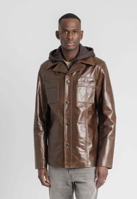 Bruine leren jack met een sluiting aan de voorkant met knopen, puntige kraag, twee borstzakken en een gladde, glanzende textuur. Draag het over een grijze hoodie.