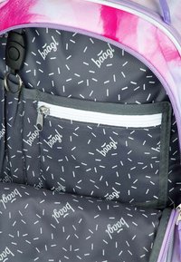 Vnitřek batohu s šedou látkou, na které jsou bílé rozptýlené čáry a text. Obsahuje uzavíratelnou kapsu a látku v odstínech fialové a růžové.