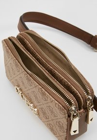 Hnědá kabelka s vzorem, se dvěma zipy, zlatými kovovými detaily a hnědým popruhem. Materiál vypadá texturovaně s logem.