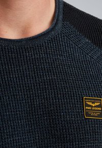 Ciemnoniebieski sweter z okrągłym dekoltem, wyróżniający się teksturowanym designem i żebrowanym wykończeniem, z czarną wszywką na ramieniu oraz żółtą łatą z logo.