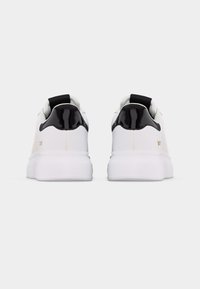 Ζευγάρι λευκά αθλητικά παπούτσια με χοντρές σόλες και μαύρες γυαλιστερές λεπτομέρειες στη φτέρνα, σε οπίσθια όψη, σε απλό φωτεινό φόντο.