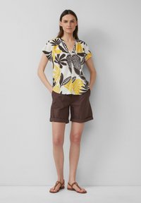 Chemise blanche à manches courtes avec imprimé de feuilles noires et jaunes, assortie avec un short marron. Les sandales ont une lanière avec un accent circulaire.