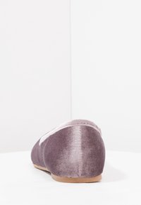 Mjuk lila sammetstuss med en ljusrosa kant och beige sula, sedd bakifrån på en vit yta.