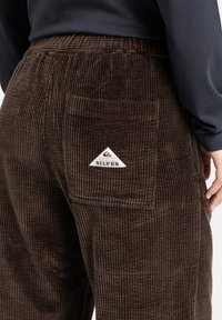 Bruine corduroy broeken met verticale ribbels, voorzien van een achterzak en een driehoekig "Quiksilver" logo patch in wit en rood.