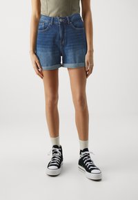 Denim šorti vidēji zilā krāsā ar saliektām malām. Iekļauj priekšējās kabatas, pogu aizdari un vieglu izbalēšanu tekstūras kontrastam. Saskaņoti ar melniem sporta apaviem.