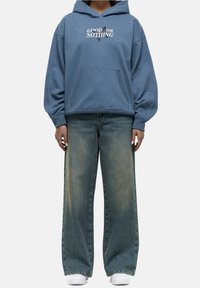 Light blue hoodie with large front pocket and printed text “GOOD FOR NOTHING.” Paired with wide-leg, faded denim jeans and white sneakers.