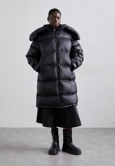 Abrigo puffer largo negro con un cuello alto, cierre de cremallera y bolsillos laterales, con secciones acolchadas y un acabado mate, combinado con una falda negra y botas.