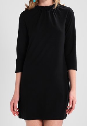 Femme portant une robe noire simple longueur genou avec manches trois-quarts et encolure froncée, debout devant un fond uni.