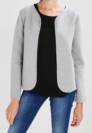 Kvinde iført en lysegrå, tekstureret åben-front jakke over en sort top og blå jeans, stående mod en ensfarvet lys baggrund.