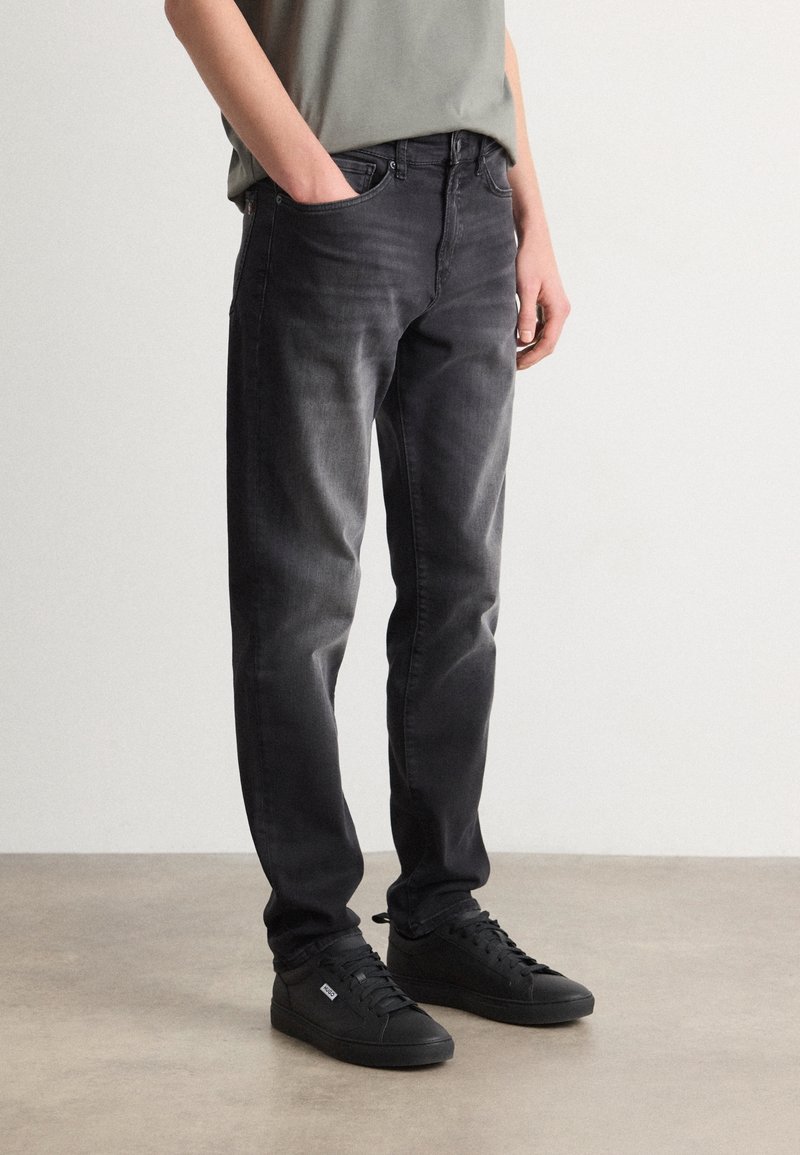 Personne portant un jean noir délavé avec les mains dans les poches et des baskets noires, debout sur un sol gris clair devant un fond uni.