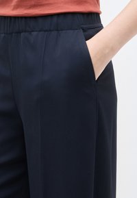 Άτομο που φοράει μπλε σκούρο παντελόνι με ελαστική μέση, με το χέρι μέσα στην πλαϊνή τσέπη, συνδυασμένο με τοπ σε απόχρωση σκουριάς.