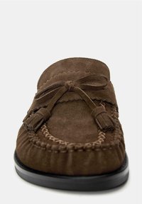 Mocassin en suede marron avec un nœud décoratif et des pampilles, surpiqûres détaillées le long de l'embout, et une semelle noire.