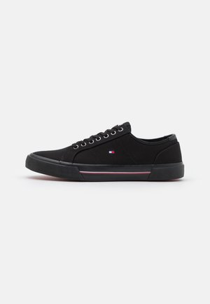 Black canvas sneakers feature a low-profile design, round toe, lace-up front, and rubber sole with a red and white stripe accent.