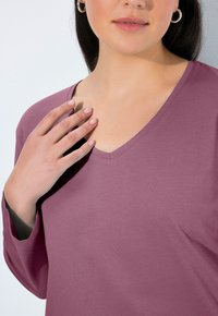 Mauve katoenen top met V-hals en lange mouwen. Kenmerkt zich door een relaxed fit en een lichte glans, met de hand van het model rustend op de halslijn.
