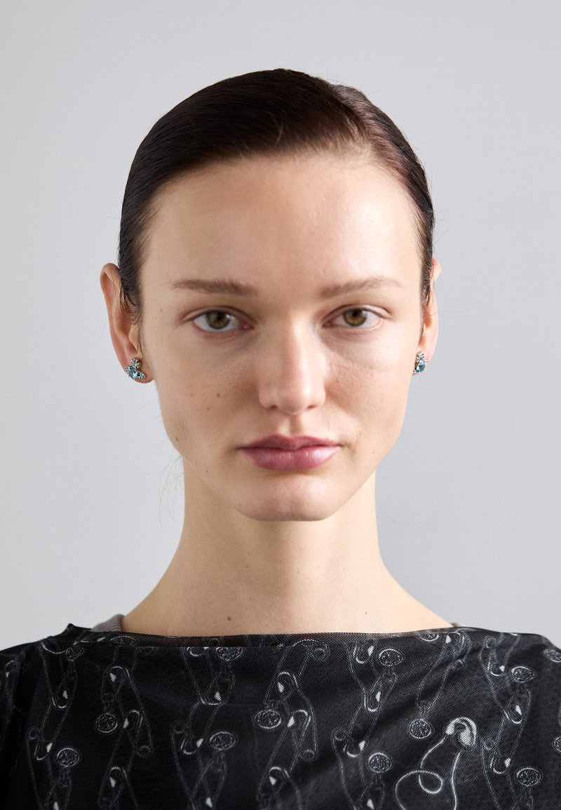 Black garment with a safety pin pattern; features a round neckline. The model wears turquoise stud earrings. Smooth skin with neutral makeup.