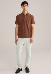 Brūns polo krekls ar klasisku apkali, īsām piedurknēm un mazu logotipu; apvienots ar gaiši zaļām biksēm un melnām mokasīnām.