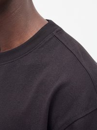 Gros plan sur l'épaule et le cou d'une personne portant un t-shirt gris foncé uni à col rond sur fond blanc.