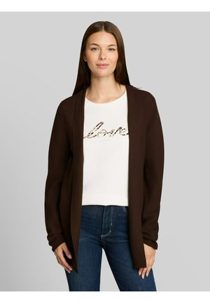 Frau trägt ein weißes T-Shirt mit dem Text "love" und eine dunkelbraune offene Strickjacke, kombiniert mit dunkelblauen Jeans, stehend vor einem schlichten Hintergrund.