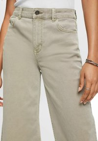 Pantalon ample couleur kaki clair en mélange de coton. Comprend des poches avant, une fermeture à bouton, et des coutures visibles le long des coutures.