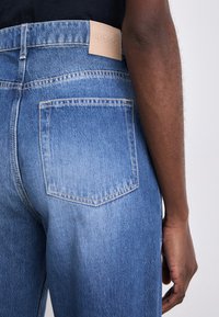 Jeans din denim de culoare albastră deschisă cu talie înaltă, având o etichetă din piele pe spate și un detaliu de buzunar cusut simplu.