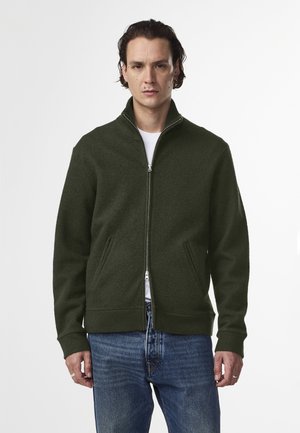 Giacca a zip verde scuro con colletto alto, due tasche frontali e polsini a coste. Tessuto testurizzato con una vestibilità casual, abbinata a jeans blu.