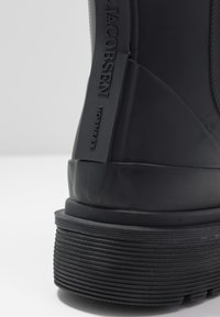 Botte en caoutchouc noire avec un dessus texturé, une semelle striée et un logo embossé sur le côté. Fini lisse avec un design durable.