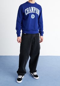 Blue sweatshirt with "CHAMPION" logo, crew neck, ribbed cuffs, paired with black baggy jeans and black and white sneakers. Simple design, casual fit.