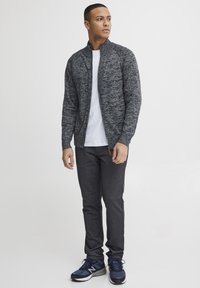 Giacca grigia a maglia con zip e colletto alto, indossata sopra una maglietta bianca, abbinata a pantaloni grigio scuro e scarpe da ginnastica blu.