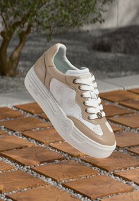 Sneaker bianca e beige con dettagli traforati, materiali in pelle e sintetici, lacci bianchi e suola in gomma, sollevata sopra una superficie piastrellata.