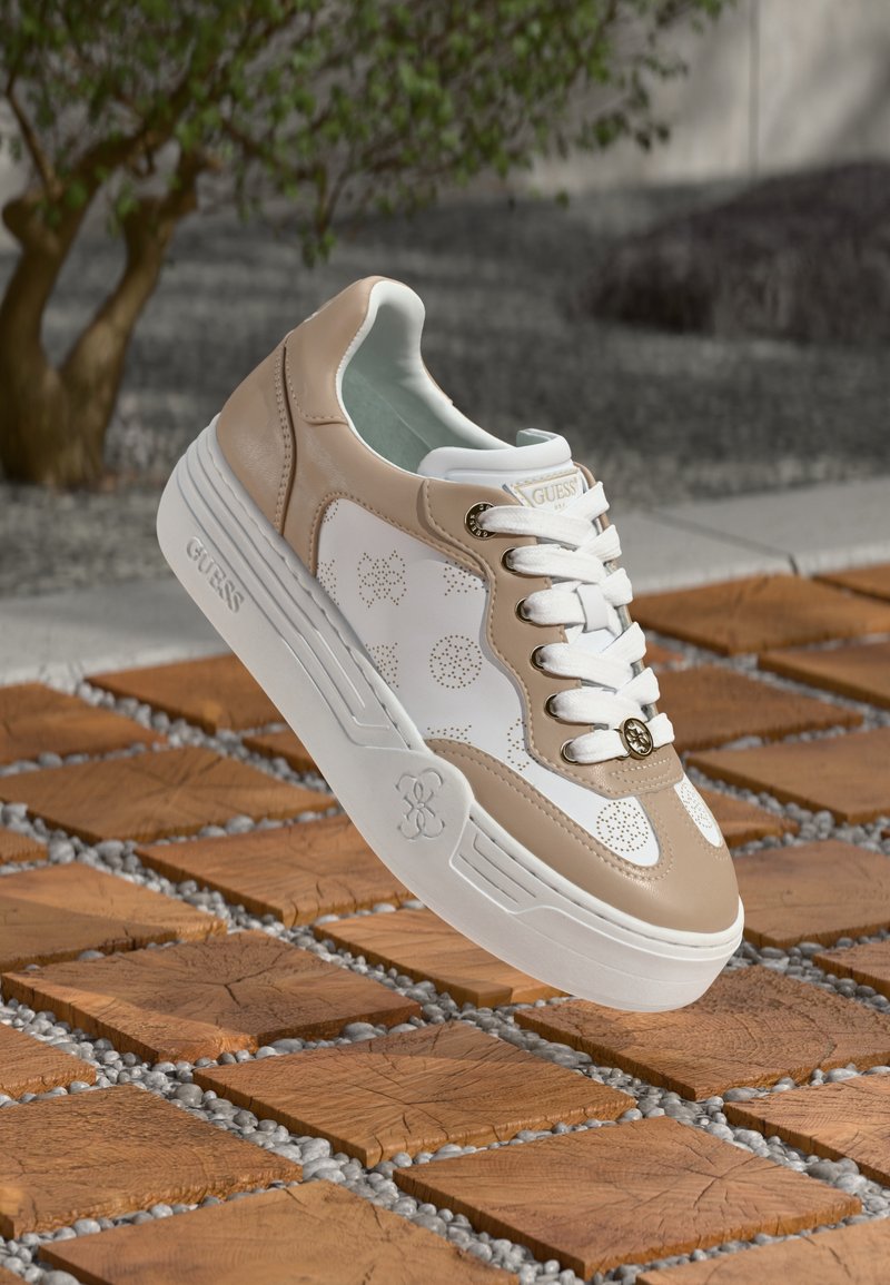Sneaker bianca e beige con dettagli traforati, materiali in pelle e sintetici, lacci bianchi e suola in gomma, sollevata sopra una superficie piastrellata.