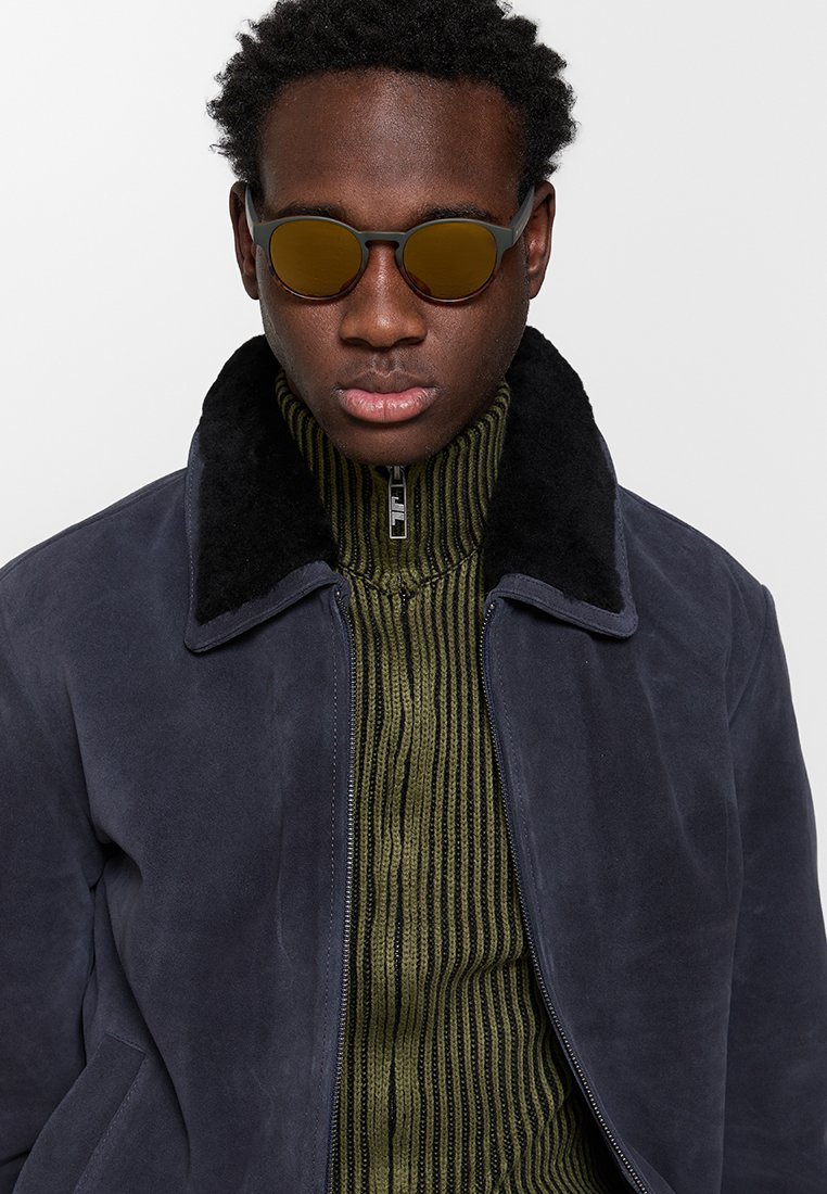 Granatowa zamszowa kurtka z czarnym futrzanym kołnierzem, noszona na oliwkowym, prążkowanym swetrze. Okrągłe okulary przeciwsłoneczne z brązowymi soczewkami.