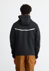 Sweatshirt preto com um bolso canguru na frente, apresentando uma faixa horizontal branca com a marca na parte superior das costas. O tecido é liso e texturizado.