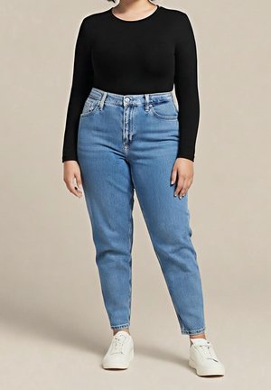 Persoon draagt een zwarte top met lange mouwen, lichtblauwe jeans met hoge taille en witte sneakers tegen een effen beige achtergrond.