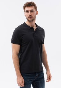 Musta polo-paita, joka on valmistettu puuvillasekoitteesta. Siinä on kauluspanta, lyhyet hihat ja hienovarainen logo rinnassa. Väri on yhdistetty denim-farkkuihin.