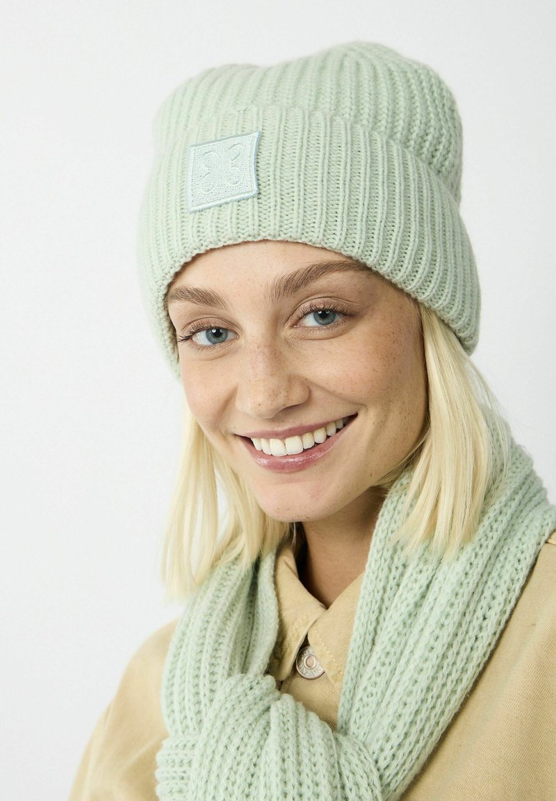 Mint green knit beanie with ribbed texture and a patch detail, paired with a matching scarf of similar texture and color.