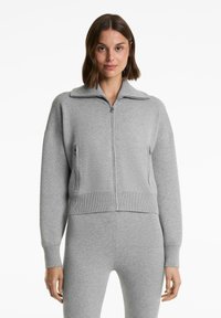 Vrouw in een lichtgrijze zip-up trui met hoge kraag en bijpassende grijze strakke broek, staand tegen een witte achtergrond.