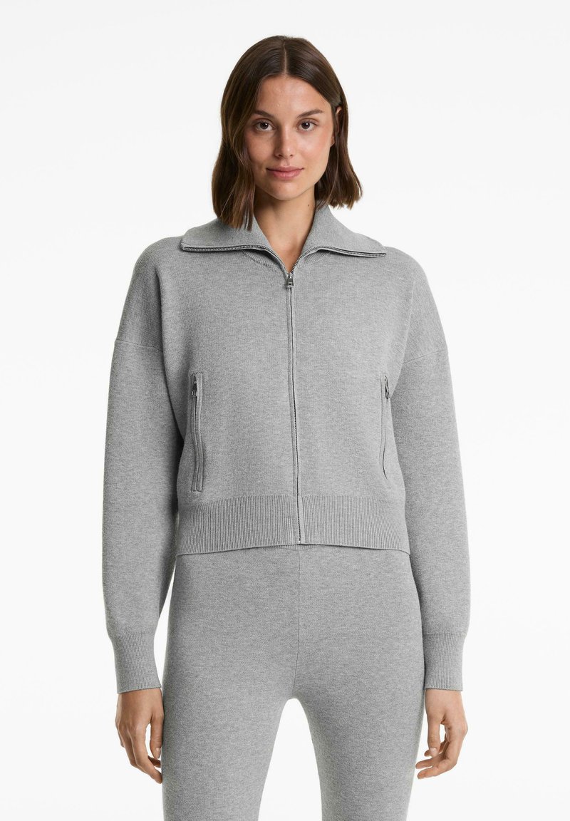 Vrouw in een lichtgrijze zip-up trui met hoge kraag en bijpassende grijze strakke broek, staand tegen een witte achtergrond.