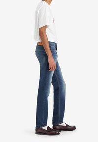 Un jean en denim bleu présente une coupe droite, des coutures contrastées et une étiquette en cuir à l'arrière. Associé à un t-shirt blanc et des mocassins.