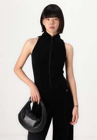 Black sleeveless jumpsuit with a high collar and front zipper. Model holds a small black leather bag with a rounded shape and soft texture.