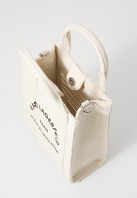 Borsa tote in tela color crema con manici corti, chiusura a scatto e scomparti interni per carte. Presenta testo stampato su un lato, design minimalista.