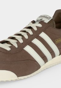 Baskets Adidas marron avec des bandes blanches, des lacets blancs et un logo marqué sur la languette et le côté, inclinées pour montrer le côté extérieur et les détails avant.