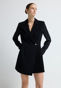 Czarna, dopasowana sukienka w formie żakietu z długimi rękawami, wyciętym kołnierzem, zapięciem na jeden guzik oraz plisowanym dołem. Gładka faktura tkaniny.