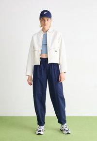 Vit kort jacka, blå figursydd topp, marinblå cargo-byxor, marinblå keps, flerfärgade sneakers, står på grön yta.