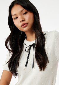 Suéter blanco de punto con cuello redondo, mangas cortas y lazo de cinta negra, adornado con acentos parecidos a perlas en toda la tela.