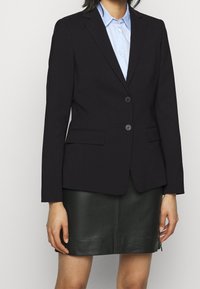 Svart skräddarsydd blazer med två knappar, slagkrage och sidofickor, parat med en ljusblå skjorta med krage och lädjerock.