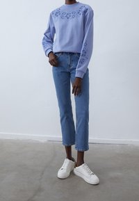 Felpa lilla con design a fiori ritagliato sulle maniche, abbinata a jeans cropped blu chiaro e sneakers bianche con dettagli dorati.
