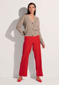 Beige gebreide cardigan met een V-hals en knopen, gecombineerd met rode wijde broekspijpen. Model draagt bijpassende rode flats. Schoon, minimalistisch achtergrond.