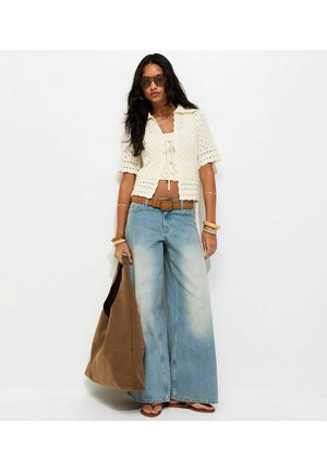 Kobieta ubrana w kremowy top na szydełku, szerokie, znoszone niebieskie dżinsy, brązowy pasek, sandały, okulary przeciwsłoneczne, bransoletki i trzymająca dużą brązową torbę na ramię.